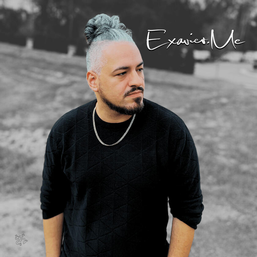 Exavier.Me Gallery Image 3 Exavier Me standing and he looks off to the side, wearing a black sweater shirt and his man-bun very present in the image.