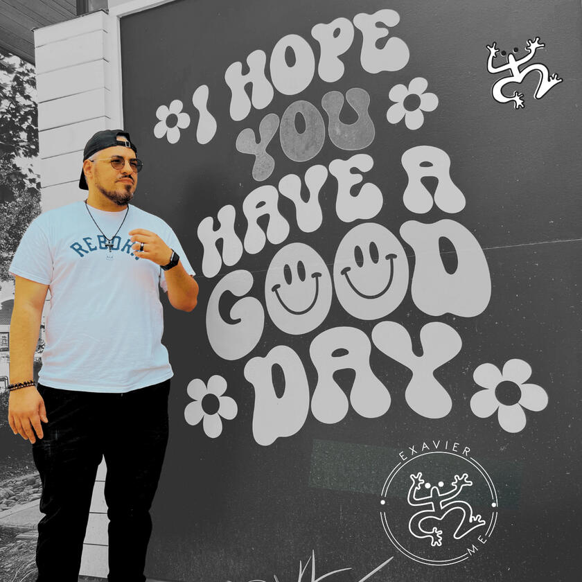 Exavier.Me Gallery Image 5 Exavier Me standing in front of a sign that reads "I hope you have a good day" while wearing a backwards snap-back cap, circular sun glasses and a T-Shirt that reads "Reborn."