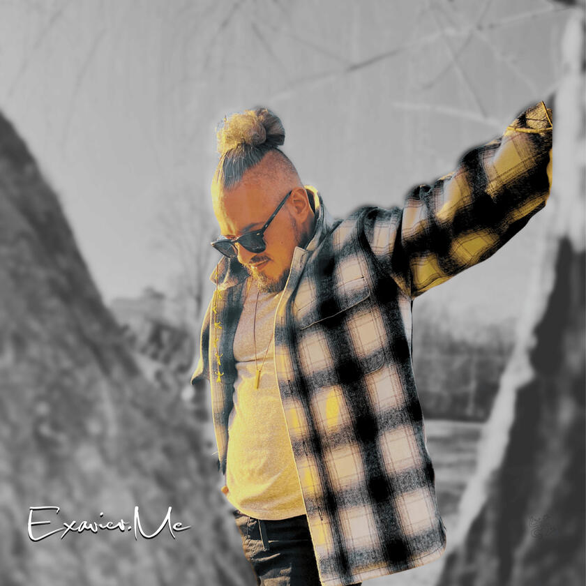 Exavier.Me Gallery Image 1 Exavier Me posing behind a tree, with a left hand leaning on a tree, the other in his pocket, as he looks down, his man bun and sunglasses are very noticeable.